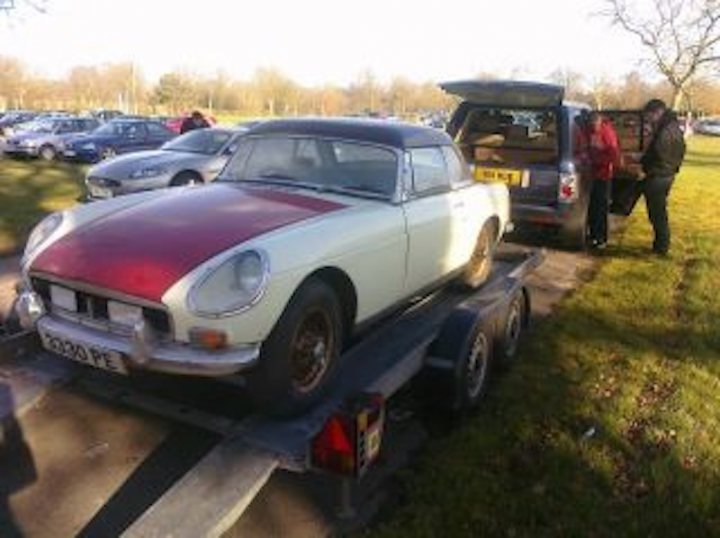 MK1 MGB body shell wanted  - Page 1 - MG - PistonHeads - In the image, there is a scene in a parking lot with several cars. The main focus is an old, white, and red two-door convertible truck. It's parked on red and white cones, suggesting that it's either being serviced or displayed for an event. The truck has a sporty appearance with its hood raised, showcasing its engine. There are two people standing near it, possibly attending to the vehicle or admiring it. The parking lot isn't empty, as there are several other cars parked in the background, adding depth to the scene.