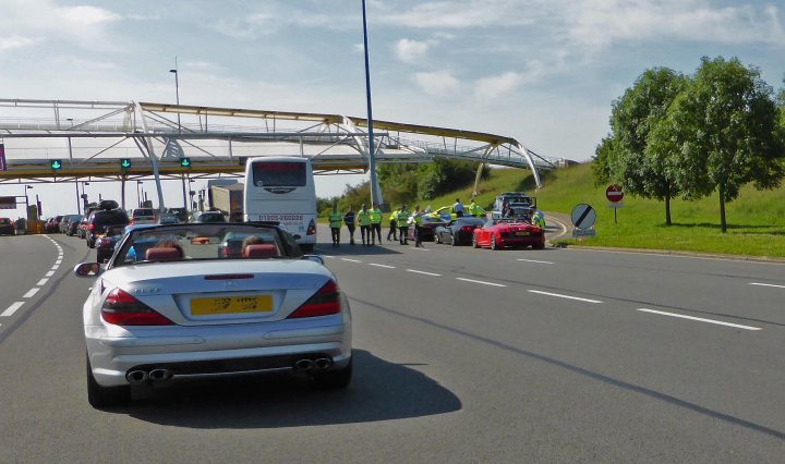 Pistonheads - The image shows a busy highway scene with heavy traffic. There are several vehicles on the road, including cars and a bus. Some of the cars are parked, and there is a person crossing the road at a designated area. The highway is lined with a metal fence and trees. Traffic lights are visible, controlling the flow of vehicles. The sky is somewhat overcast, suggesting it might be a cloudy day.