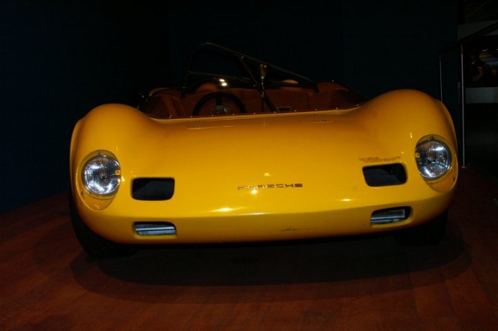 Pistonheads - The image shows a vibrant yellow sports car, possibly a Porsche, elegantly displayed in an art gallery. The vehicle's sleek design, highlighted by the prominent headlights and the convertible top, exudes a sense of luxury and speed. It's parked on a wooden floor, which contrasts with its color, providing a unique display. The gallery's overall setting and the play of light and shadow contribute to the artful presentation of the car.