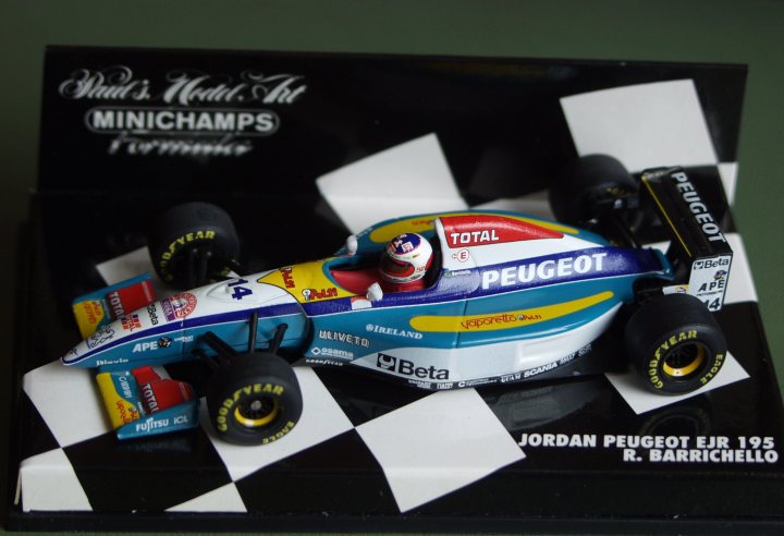 Collection Pistonheads - This image shows a close-up of a model car on a checkered racing display base. The car is a toy representation of a Formula 1 racing car, designed to resemble the livery and branding of the Peugeot team. The model car is predominantly blue and white, with accents of red, yellow, and black, and features the word "PEUGEOT" prominently displayed on its side. The base of the model car displays the name "R.BARRICHELLO," likely referring to the driver it is meant to represent, and other racing-related graphics. The overall scene is a staged representation capturing the theme of a miniature racecar display.