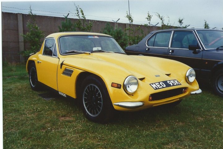 A red car is parked on the grass - Pistonheads - The image shows a vintage yellow sports car parked in a grassy area with a brick wall in the background. Another car with a black exterior is partially visible on the right side of the frame. The sports car features a distinctive design with a large grille, sporty vents on the hood, and a hardtop. There's a BE0 95K license plate on the front of the car, suggesting it may be a special edition or part of a club. This vehicle has a classic silhouette and appears to be in good condition, potentially a collector's item.