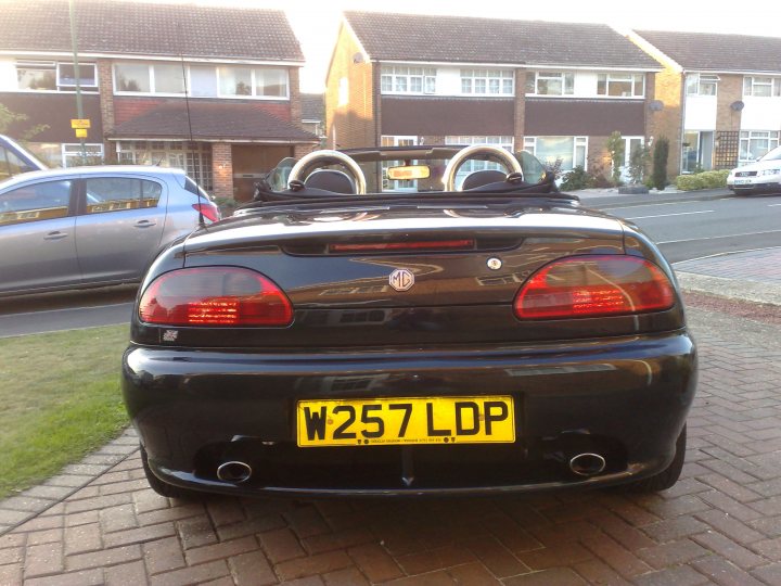 Pistonheads - The image showcases a sleek, black sports car with a distinctive yellow and black license plate. The license plate reads "W25 7LDP." The car is parked on what appears to be a brick driveway, adjacent to a residential street where other vehicles are visible. The setting suggests a residential area with a house visible in the background. The sunlight in the image gives a warm, golden glow to the scene.