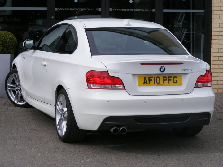 Pistonheads - The image displays a white BMW coupe parked in what appears to be a dealership lot. The vehicle is parked in front of large sliding glass doors, suggesting the presence of a dealership or service center in the background. The BMW model is visible above the license plate, which reads "AF10 PGP." The scene is taken during daylight, as indicated by the shadows and the bright visible sky.