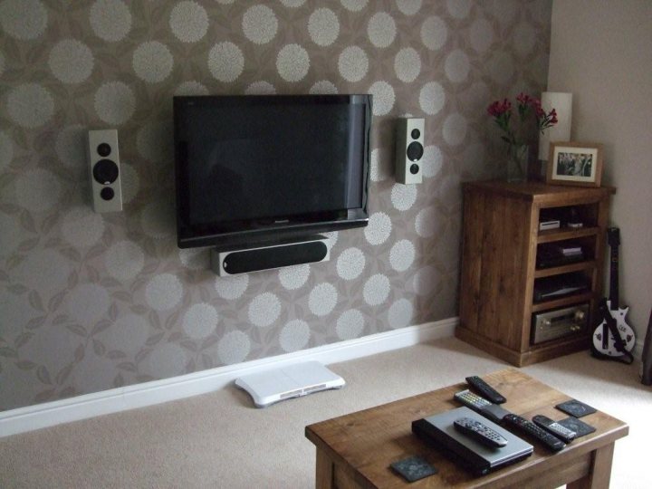 Mounting Pistonheads Wall - The image depicts a cozy living room setup. Against a textured wall with a floral pattern, a flat-screen television is mounted above a remote control, allowing for easy access to the TV's functions. A wooden cabinet sits in the corner, adorned with flowers and other decorative items. The wooden table is topped with various remote controls for different devices in the living room, positioned in front of the cabinet for convenient access. There is a glimpse of a guitar leaning against a cabinet on the right side of the photo. The overall ambiance of the room is warm and inviting, suggesting a space designed for relaxation and entertainment.