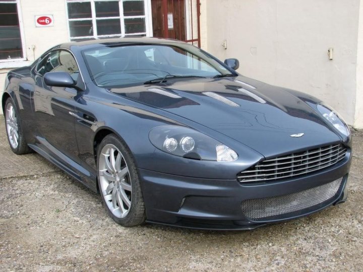 Pistonheads - The image shows a dark-colored, convertible sports car parked on a gravel driveway. The car features a sleek design with a distinctive front grille, headlights, and alloy wheels. The car appears to be of a premium or luxury category. In the background, there is a hint of a residential building, but the focus is clearly on the car. The composition of the image suggests it might be for sale or display purposes, given the pristine condition and setting.