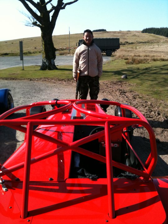 Wales Blat Pistonheads - In this image, a man is standing in front of a vibrant red sports car with a convertible top. The car is prominently displayed in the foreground of the picture. The man is holding a gun and seems to be posing proudly next to the car. Behind him, there's a serene landscape that includes a lush green field and distant mountains under a blue, sunny sky. The open terrain and the clear blue sky suggest a peaceful day.