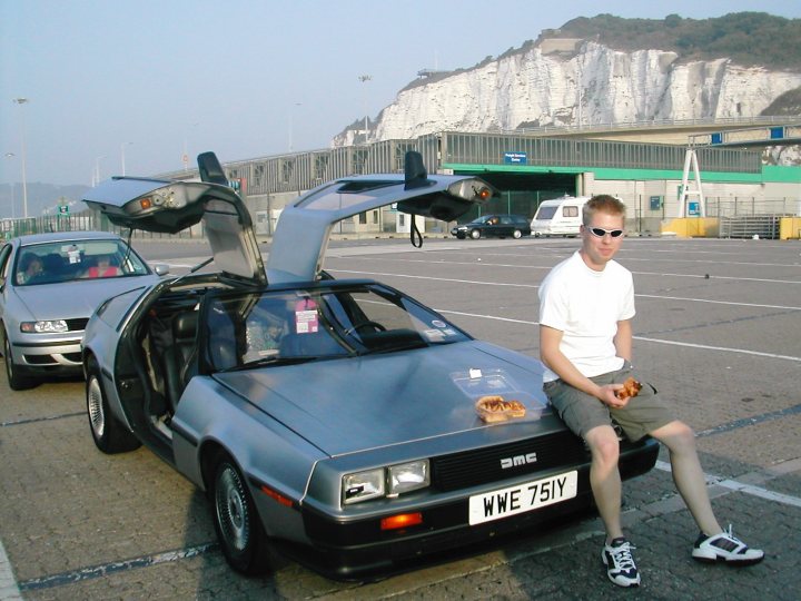 Your 'No longer with you' pride and joy... - Page 1 - Readers' Cars - PistonHeads - In the image, a man is seated in a vintage car with the ajar doors, which sets it apart from the others. The car he's occupying is a DeLorean, a unique car characterized by a distinctive stainless steel exterior and gull-wing doors. The backdrop includes a parking area and chalk-white cliffs, suggesting a scenic location far from a run-down area.