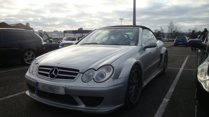 Oxford Sunday Service Pistonheads - In the image, a silver Mercedes-Benz sports car is parked in a large parking lot under a cloudy sky. The car is parked next to a white line, and a lifeguard tower is visible in the background. The car's design is sleek and its bodywork has a few dents and scratches on it. A person is visible walking near the car.