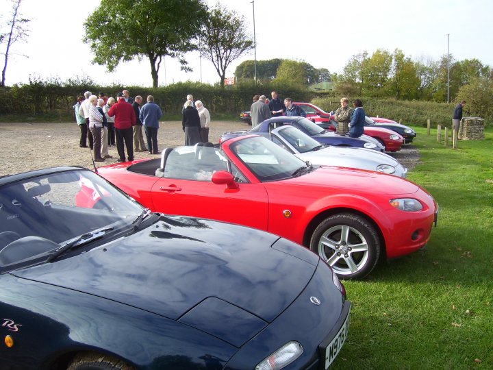 Region Owners Pistonheads Western October - In the image, there's a group of people gathered in a field, standing around various types of cars parked in a line. They appear to be inspecting or engaging in conversation about the vehicles. The cars themselves are diverse, including both convertibles and fixed-top models, all exhibiting a range of colors and designs. The field they are in is expansive, stretching out to meet the horizons and providing a spacious backdrop for this gathering. The sky overhead is overcast, suggesting that it might be a cooler day.