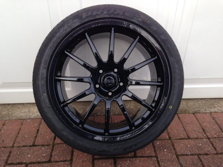 Alloy wheel referb, Newcastle area - Page 1 - North East - PistonHeads - In the image, a single black car wheel is resting on a brick floor. The wheel seems to be in good condition with no obvious signs of wear or damage. It has a five-pointed star hub bolt pattern, a common design for vehicles like BMWs or Audis. The wheel is positioned close to the bottom of the image, providing a clear view of its detail. A corner of a white door can be seen in the background, suggesting the image might have been taken near a building or a parked vehicle.