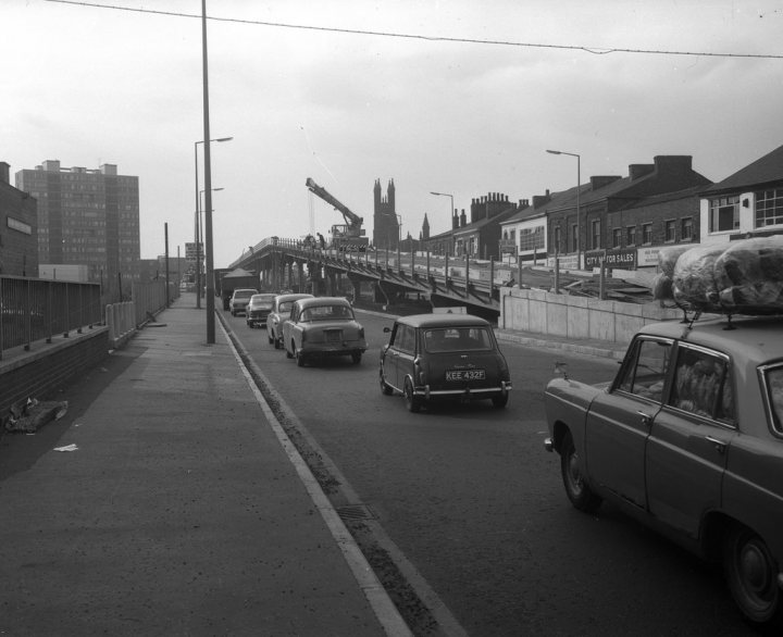 How about a 'period' classics pictures thread - Page 500 - Classic Cars and Yesterday's Heroes - PistonHeads - This is a black and white photo capturing a bustling cityscape. The street is filled with a variety of cars, including an old-model hatchback and an even older car, suggesting a busy city traffic scenario. Buildings of varying heights, possibly including a cathedral, line the street, adding architectural interest to the scene. A walkway spans the road, providing a pedestrian crossing. A crane is visible in the background, indicating ongoing construction or development activity.