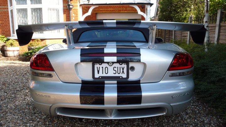 Got her out  - Page 1 - Vipers - PistonHeads - The image shows a rear view of a silver sports car, which appears to be a V10 SUV given the text on the license plate. The vehicle's rear quarter panel is prominently visible, featuring dark and light silver diagonal stripes. It's parked on a gravel driveway that leads to a brick wall, and there's a house and vegetation in the background. The license plate is distinctive and appears to be personalized.