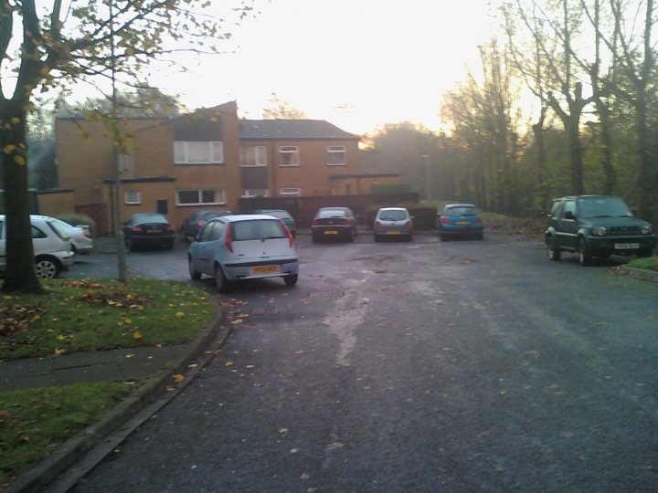 Pistonheads Parking Bad Thread - The photo captures a residential street scene. Several cars are parked on the street, with their bodies facing the camera. The vehicles come in shades of white and blue and vary in model and size. A few cars are closer to the camera, while others are parked further back, indicating a long street. To the side of the street, there are trees with leaves on the ground, suggesting either a fall or spring season. The sky is overcast, and there are no people visible in the scene.