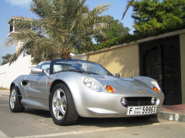 A classic car is parked in a parking lot - Pistonheads - The image features a modern, grey two-door sports car parked in front of a low wall adorned with potted plants. The car has a sleek design with clean lines and alloy wheels, carrying a license plate that reads "F 59636". Large, leafy palm trees frame the scene, adding to the exotic ambiance. The sky is partly cloudy, suggesting a warm, pleasant climate.