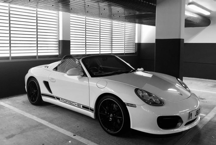 Boxster Spyder - Page 472 - Boxster/Cayman - PistonHeads - The image shows a white sports car parked in an indoor parking area. The vehicle is sleek, with a design that suggests it's made for speed and luxury. It has distinctive features such as large alloy wheels, a long bonnet, and aerodynamic curves. The license plate of the car is clearly visible. The car is positioned at an angle to the viewer, giving a profile view of the vehicle. The background is dimly lit, indicating that it may be evening or located in a garage or indoor parking facility.