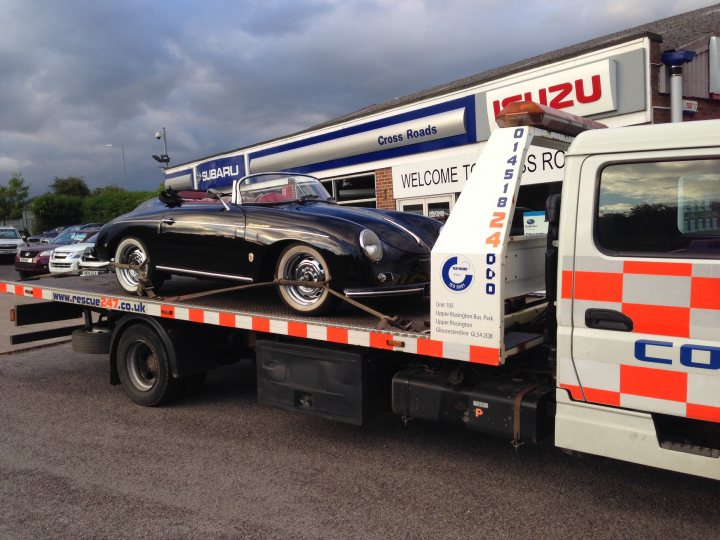 So what have you done with your Aston today? - Page 123 - Aston Martin - PistonHeads - The image showcases a vintage black sports car being towed on a flatbed truck. The car, with its sleek design and large wheels, is the centerpiece of the scene. The vehicle appears to be in pristine condition, suggesting a cherished or valuable possession. The flatbed truck, which is predominantly white with orange stripes and the word "Code" written on the side, is used to transport the car from a location suggested by the "Izu Road" sign in the background. The scene takes place during the day under an overcast sky.