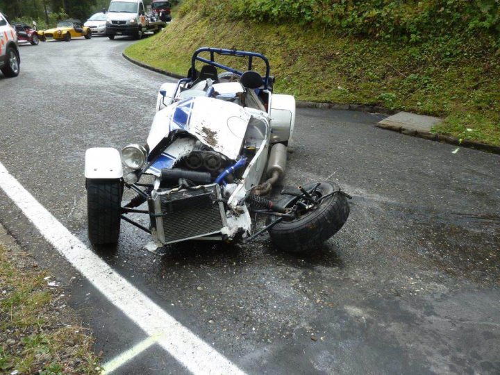 Caterham crash repair - Page 1 - Caterham - PistonHeads - The image depicts a chaotic scene on a road where a single-engine racing car has flipped and is resting upside down. The car's body is dislodged from its frame, revealing its engine and suspension. The surrounding area is scattered with debris, and there are several vehicles parked on the side. The road appears to be wet, possibly due to recent rain, and the grassy embankment on one side is also spotted with chunks of concrete.