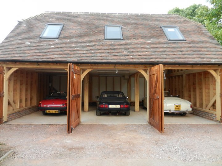 Out in the cold - Page 2 - S Series - PistonHeads - The image shows a traditional wooden garage housing a variety of cars. There is a garage door open, revealing a red car parked inside, and revealing another car parked inside a neighboring garage. Through the adjacent door, a second garage space with a black car parked is visible. The garage has a peaked roof and is open to the elements. The setting appears to be a residential area, possibly a driveway, given the presence of the garage and partially visible driveway stairs leading to the garage door.