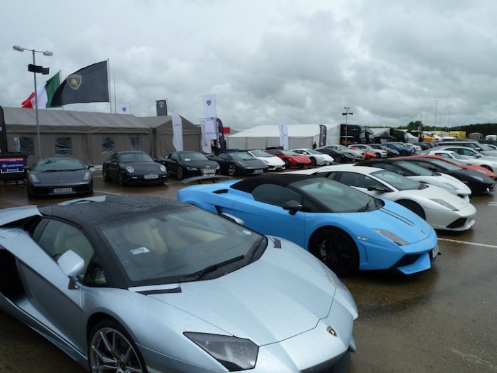 Lamborghini Super Trofeo and Blancpain GT  - Page 1 - Lamborghini Classics - PistonHeads - The image depicts a scene from an outdoor auto show. The lot is filled with a variety of cars, symbolizing different brands and models. Dominating the foreground are two luxury cars. On the left, there's a silver sports car, sleek and modern in design. Adjacent to it on the right, a vibrant blue sports car showcases its sleek lines and elegant details. The cars are parked on concrete, and behind them, several tents stand erect, possibly housing more exhibits or serving as hospitality areas. Above, a cloudy sky casts a muted light over the entire scene, enhancing the colors of the cars and providing a somber backdrop to the vibrant display of automotive engineering.