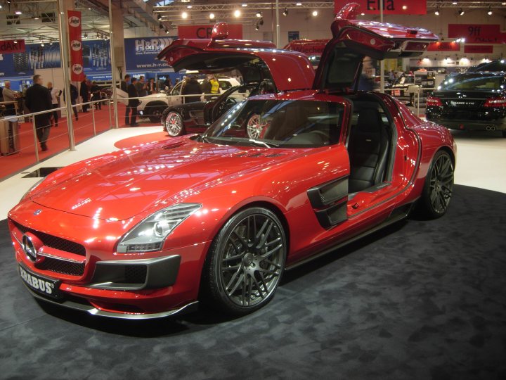 Pistonheads Highlights Essen - The image portrays a sleek, black roofed red sports car exhibited at an auto show. The car is parked indoors in a spacious showroom or exhibition hall, which has a carpeted floor. The white walls and multiple banners adorned with company logos suggest a professional, commercial setting. The shock absorption is blown out, revealing the suspension system and the ignition is open, revealing the engine, indicative of testing or demonstration.