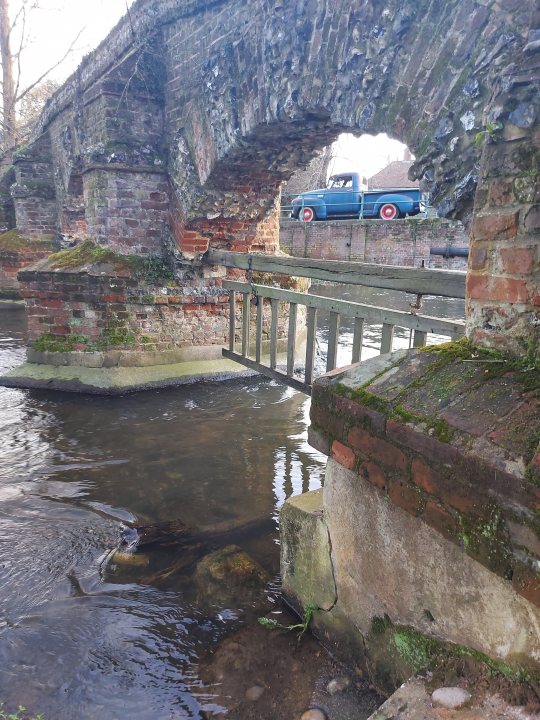 Pistonheads - The image shows a picturesque scene of an old brick bridge with an archway. A small, blue car is crossing the bridge, which appears to be in good condition despite its age. The bridge is nestled amidst a lush green landscape, possibly indicating that it's located near a body of water. Surrounding the bridge are trees and shrubbery, adding to the serene atmosphere. The bridge and car are the main focus of the image, with their placement suggesting they are moving downstream along the river.