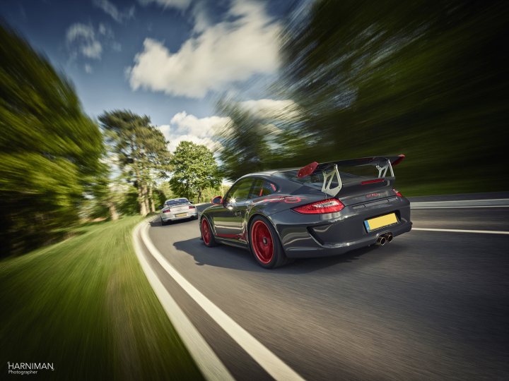 The Beautiful Porsche Picture - Page 4 - Porsche General - PistonHeads - The image captures a dynamic scene on a winding road. At the center, a majestic black sports car with a large spoiler and red wheels is driving around a bend, showcasing its power and agility. There are two other vehicles in the frame, accompanying the sports car on their own journey. The road itself is lined with lush green trees, adding a sense of serenity to the otherwise high-speed action. The background is a blur of blue and white, likely the sky, implying the setup for a car advertisement or racing gallery.