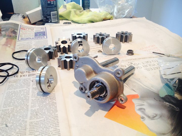 A stack of books on a table with a clock - Pistonheads - The image is a detailed close-up of a workspace, and it features a collection of car parts, which include various metal pieces, potentially gears and bearings, scattered across a newspaper-covered table. The camera angle is from directly above the central section of the table, emphasizing the mechanical elements. On the right side, there's a photo of a cartoon-style female figure. In the background, there's a glimpse of a green rag, suggesting an ongoing work or cleaning process. The layout and the items indicate a mechanic or automotive enthusiast's environment.