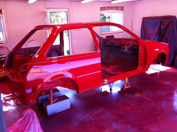 Fiesta RS Turbo Restoration - Page 7 - Ford - PistonHeads - The image shows an open car door in a car painting or repair workshop. The car is primarily painted in a vibrant red color. The paint appears to be drying on the floor and walls around the car, indicating that the body was likely painted recently. The interior visibility is limited due to the open door, but the general space seems ample, typical of a workshop area. The car is placed on a floor that has some remnants of other car parts or materials on it, implying a working environment.