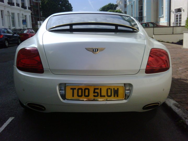 Pistonheads - The image features a silver luxury car parked on a road adjacent to a sidewalk. The car is a converted Bentley TGV, identifiable by the distinctive badge on its trunk. The design of the car is sleek and elegant, with chrome details and shiny paint reflecting its environment. On the license plate, the phrase "Too Slow" is written. The cityscape in the background includes high-rise buildings, suggesting that the car is parked in an urban area. The surroundings are characterized by a mix of residential and commercial architecture.