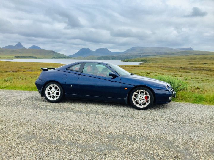 Let's see your Alfa Romeos! - Page 114 - Alfa Romeo, Fiat & Lancia - PistonHeads - The image showcases a blue sports car parked on the side of a gravel road. The car is positioned in the foreground, with a backdrop that features a tranquil body of water surrounded by hills under an overcast sky. The vehicle appears to be in good condition, with the shiny blue paint gleaming under the soft light. There are no people visible in the image, focusing the viewer's attention solely on the car and its natural surroundings.