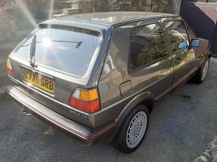 1986 Golf GTi - Page 1 - Readers' Cars - PistonHeads - This image features a vintage car, likely from the 1980s or earlier, parked on a concrete surface. The car has a metallic silver finish and appears to be in good condition. It's equipped with a sunroof and a roof rack. There are two visible license plates, one of which reads "76KJ" indicating it could be a UK-registered vehicle. In the background, there's a structure that looks like a building wall, contributing to an urban setting. The overall impression is that of a well-kept classic car on display in an outdoor space.