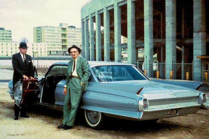 A 'period' classics pictures thread (Mk III) - Page 41 - Classic Cars and Yesterday's Heroes - PistonHeads UK - This image features a vintage scene where two men are standing next to a classic car, which appears to be an older model with distinctive design elements. One man is positioned in front of the vehicle while the other stands behind it. The car is parked on a concrete surface in what looks like an urban environment, possibly near a parking structure or a similar industrial structure. The setting suggests a moment captured from the past, given the attire and style of the men and the car. There's no text present in the image.