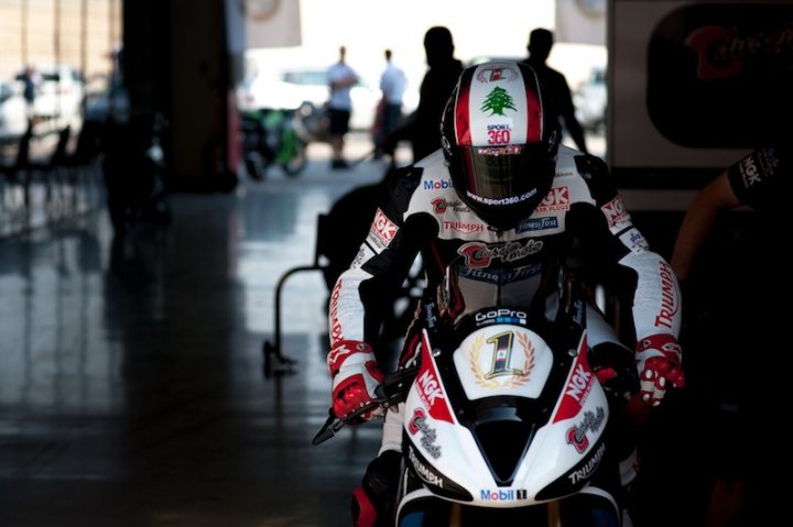 Day Race this Friday - Page 1 - Middle East - PistonHeads - The image captures an indoor scene where a motorcycle racing suit is prominently displayed. The suit is primarily white with red and black accents, and it's partially zipped up. The text and sponsor logos on the suit are in English, but without a clear view, the specific words are not identifiable. There are a few people in the background, one of whom is holding a bicycle, but they are not the focus of the image. The setting seems to be a commercial or retail space, as indicated by the neatly arranged items and the presence of logoed objects. The perspective of the photo is from a low angle, looking up at the suit and the people, which adds to the grandeur and importance of the displayed motorcycle suit.