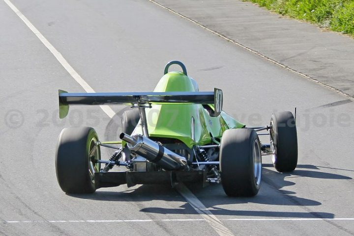 Pistonheads - The image shows a high-performance green race car on a tarmac road during what appears to be a race. The vehicle's bodywork and tubular chassis are prominent features, indicative of complex aerodynamics and lightweight materials typically used in this type of racing. The front and rear wings are designed to improve downforce and optimize performance on the track. The setting suggests a professional racing scenario, with clear lines on the pavement indicating a race circuit.