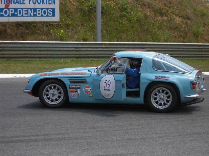 Early TVR Pictures - Page 126 - Classics - PistonHeads - The image shows a vintage car on a race track, with its roof removed and its nose pointed slightly downwards. The car is painted in a vibrant shade of blue and is gliding along a smooth black surface. Behind the car, there is a grassy area and a street sign partially visible. The track has a backdrop that features a green tree and shrubbery, implying a natural enclosure for the racetrack.