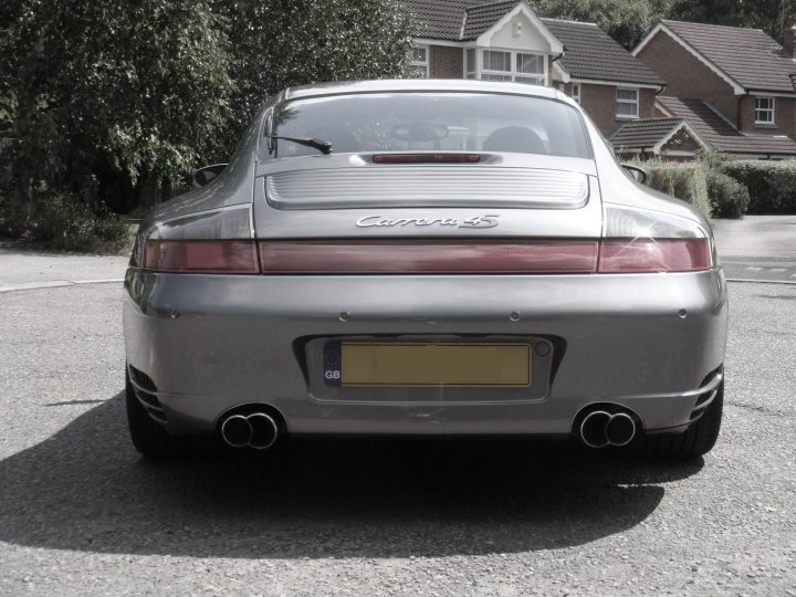 show off your porsche - Page 2 - Porsche General - PistonHeads - The image showcases the back view of a silver Porsche sports car parked on a paved street. The car is equipped with dual tailpipes and a license plate that reads "GB." The vehicle is sleek and polished, reflecting a shiny surface that catches the eye. The car is confined by a shadow cast by the tall houses in the background, creating a contrast between the urban setting and the car's sports design.