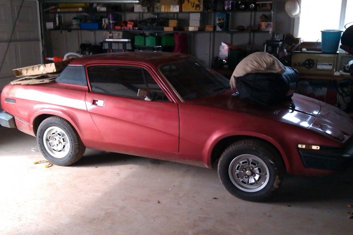 TR7 advice please - Page 1 - Triumph - PistonHeads - This image depicts a vintage red car inside a garage, surrounded by various tools and equipment. The car appears to be a classic model, possibly a Datsun or similar Japanese vehicle, indicated by its fairing and pop-up headlights. The interior rear driver side window is open, and there is a pile of cardboard boxes and papers stacked inside the car on the seat. The garage is filled with a cluttered arrangement of items on shelves behind the car, suggesting an active workspace or hobbyist environment. The overall scene conveys a sense of nostalgia and the passion for vintage vehicles.