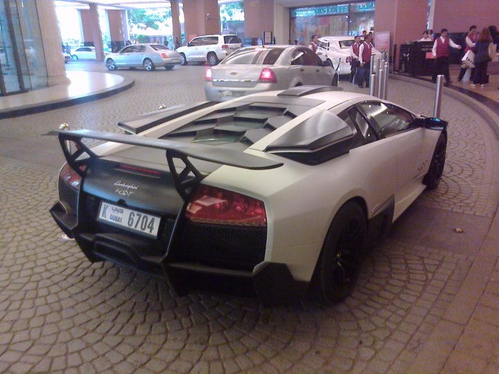 Pistonheads Supercars Spotted Rarities - The image features a luxurious, white sports car with a large rear spoiler and a black front spoiler. The car's license plate displays the number "6704". The setting appears to be a multi-story parking garage, with several other vehicles visible in the background. The vehicle is parked on a level that has a distinctive patterned floor.