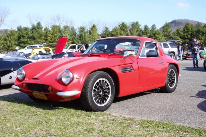 Early TVR Pictures - Page 73 - Classics - PistonHeads - The image shows a vibrant red classic convertible car parked in a gravel area with grass and multiple people in the background. There are trees giving a lush surrounding to the parking space. The car is positioned towards the viewer's perspective, suggesting it's the main focus of the photo. The lighting is bright, possibly due to the time of day or weather conditions, casting a clear and bright shadow of the car on the ground. The overall setting appears to be an outdoor event or gathering with numerous cars and individuals present.