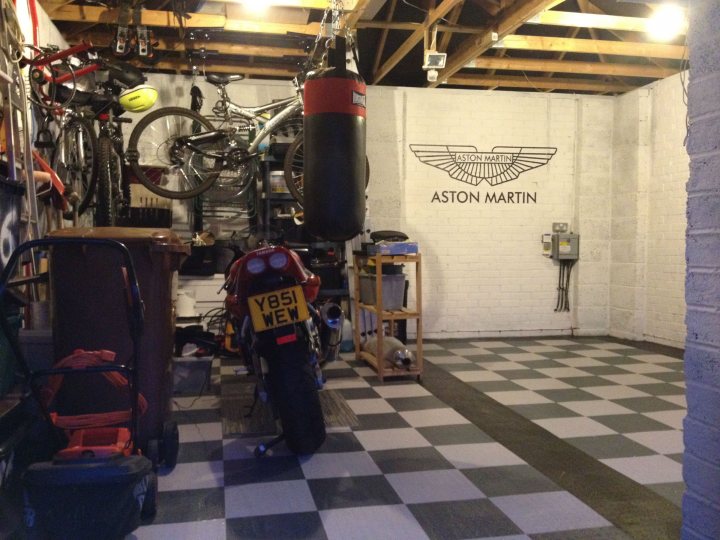 Garage Queens - Page 9 - Aston Martin - PistonHeads - The image shows an indoor space that appears to be a garage or a workshop. The main feature is a vintage motorcycle with the license plate "EYSIWE". Above the motorcycle, there is a hanging punching bag, suggesting that this space is also used for training or exercise, possibly for a martial arts enthusiast. The room has a rustic look with a wooden beam ceiling and a brick floor. On one side of the room, there is a collection of bicycles hanging from the ceiling, and there are various tools and equipment scattered around the space. The walls are adorned with the Aston Martin logo, indicating a possible interest in luxury vehicles.