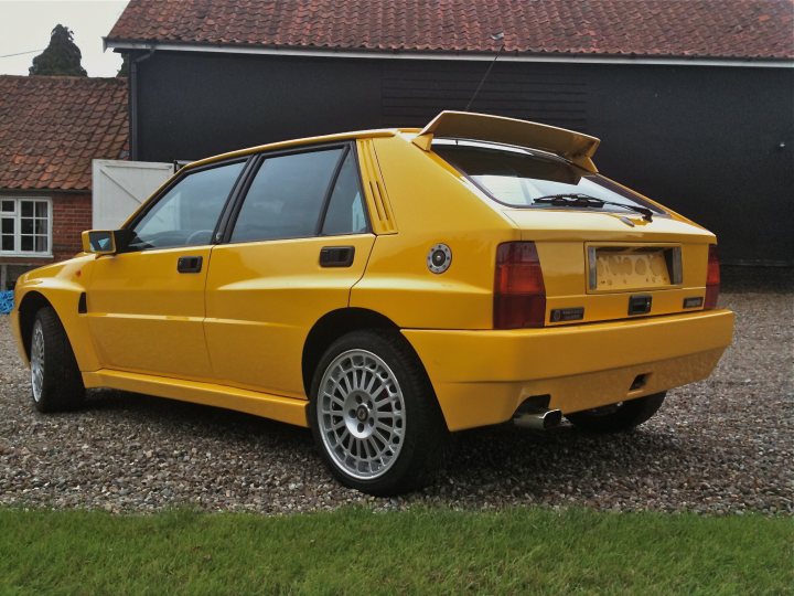 Pistonheads Endulgence - The image shows a yellow coupe parked on a gravel driveway in front of a house with a brick facade. The vehicle has a distinctive rear design with a wing spanning across its body. The moment captured in the photograph suggests it's either the morning or afternoon due to the soft, natural lighting. The coupe has alloy wheels and the license plate is clearly visible, adding a certain authenticity to the scene. The overall style of the car is reminiscent of classic sporting models.