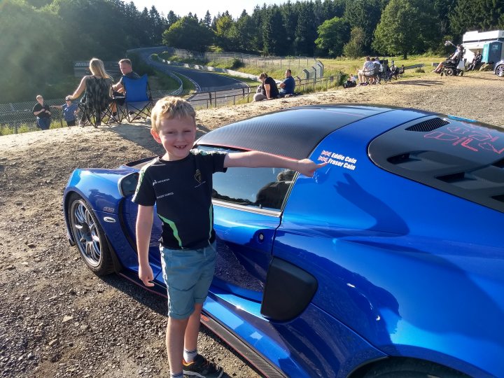 New arrival. Exige 410 Sport - Page 18 - Elise/Exige/Europa/340R - PistonHeads UK - In the image, a young boy is proudly standing next to his father's blue sports car. The car is parked on gravel, with a clear sky above and trees in the background. They seem to be at an event, as there are other cars visible in the distance. The boy is pointing to the hood of the car, perhaps showing off its features or explaining something about it to his father. It's a moment filled with excitement and pride for both the child and the adult.