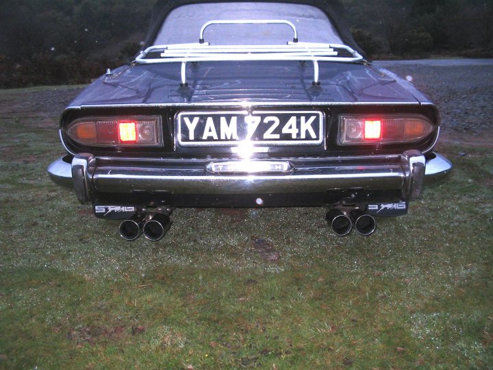 Winter Work Pistonheads - This image is a photograph of the rear end of an old-model car, which seems to be a rotary engine vehicle given the distinctive badge on the trunk lid. The car's license plate is clearly visible, indicating "YAM." The vehicle is parked in a grassy area, with a significant stocking showing underneath, revealing exhaust pipes. There is a slight reflection on the vehicle's side, possibly from a nearby water source or recent rain.