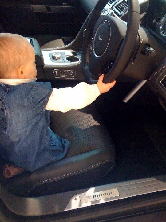 What goes with an Aston ? - Page 1 - Aston Martin - PistonHeads - The image captures a cute moment of a young child sitting in the driver's seat of a luxury car. The child, dressed in a white long-sleeved shirt and blue jeans, is observed wearing a seat belt. The car's center console houses various controls and buttons, which are clearly visible within the child's reach. The perspective of the image, taken from above the child, provides an engaging look at the scene, emphasizing the child's size relative to the car's interior.