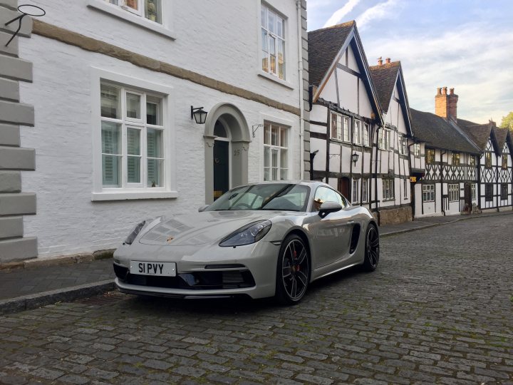 Show us pictures of your 718 - Page 1 - Boxster/Cayman - PistonHeads - In the foreground, a silver Porsche sports car is parked on a cobblestone street. The car's shiny exterior reflects the light, suggesting it's a sunny day. It's positioned in front of an older white house with a pointed archway, which features two shuttered windows and a hanging light fixture on the left side. To the right of the image, there's a larger, traditional-style white building with black beams or shutters. In the background, partially obscured by an image frame, there is a glimpse of more historic architectural styles.