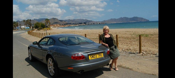 A man standing next to a parked car - The image is a photograph of a person standing beside a parked car. The individual appears to be a woman, with her back turned towards the camera. She is wearing a short-sleeve blouse and dark pants, and is holding what looks like a small handbag or purse. The car is a convertible sports vehicle, likely a BMW given its design. It's an overcast day and the background suggests a coastal town with buildings along the beachfront. There are no visible texts in the image.