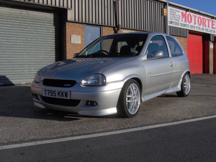 Trying to find my old car, have you seen it? - Page 1 - VX - PistonHeads - The image shows a silver Toyota hatchback car parked in a lot with a metal garage in the background. The car is facing the viewer, and the license plate starts with the number 795. The scene appears to be on a sunny day, and there's a building with a red door and a sign that reads "MOTORTECH" in the background, suggesting a garage or repair shop. The ground is covered with gray gravel, and the surrounding environment is rather industrial, with the focus on the car itself.