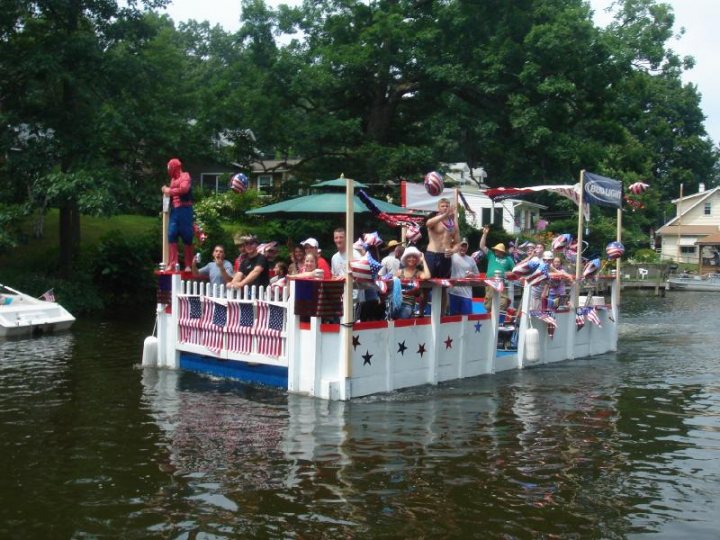 Jiggyziggy2 - The image captures a joyful scene on a body of water, possibly a river or lake. A festive, white boat with an American flag on its side is in motion, filled with numerous people. Some passengers are sitting on the railings of the boat, waving and enjoying the atmosphere. There are other boats visible in the background. The backdrop reveals the lush greenery of a wooded area, suggesting a natural, outdoor setting. This is an image during a bear celebration or event, identifiable by the multitude of small American flags adorning the boat and the celebratory mood amongst the passengers.