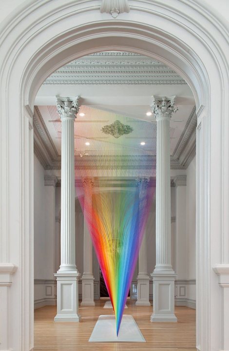 A vase with flowers in it sitting on a table - This is a photograph of an indoor space that resembles a museum or gallery. The main focus of the image is a large, rainbow-colored sculpture composed of vertical strips. The structure is set against a backdrop that includes classical architectural elements such as a columned archway and what appears to be a high ceiling with white plasterwork. The room has white walls and a wooden floor, creating an elegant and bright atmosphere. There are no visible texts or distinctive marks in the image.