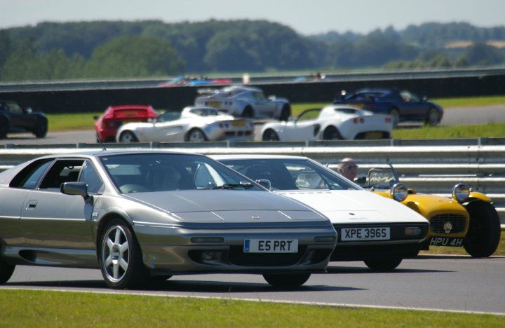 Snetterton Lotus Festival 25/26 June - Page 1 - General Lotus Stuff - PistonHeads - The image is a vibrant depiction of a car racing event. There are various rows of race cars, ready to speed off around the track. These cars, distinguishable by their prominently displayed number plates, are captured in a blurred motion likely during a race. The track itself is lined with barriers, ensuring the safety of both the racers and the spectators. The backdrop presents a lush green field, creating a striking contrast with the high-energy scene unfolding within the racing circuit.